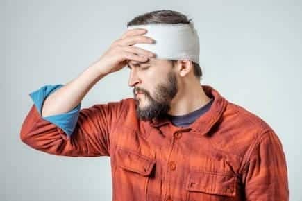 A bearded man in an orange shirt, with a bandaged head, touches his forehead with a pained expression, embodying the silent struggle that often follows the main causes of traumatic brain injury.