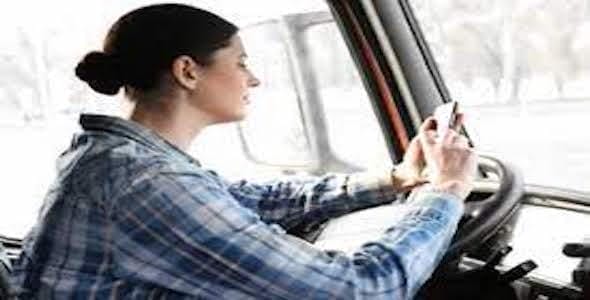 A woman in a plaid shirt is sitting in a vehicle, glancing at her cell phone while holding the steering wheel, mindful of Michigan's strict texting laws.