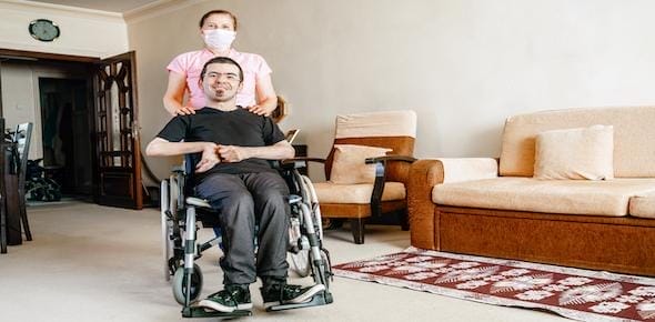 A person seated in a wheelchair, a brain injury survivor, is accompanied by another standing behind them in a cozy living room with beige furniture and a rug.