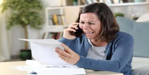 A woman is sitting on a couch, holding a phone to her ear and looking at a piece of paper with a distressed expression, likely dealing with the aftermath of a Michigan car accident and struggling through the complexities of auto insurance claims.