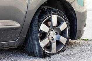 A car with a flat tire is parked on gravel, its sidewall damaged and torn from an apparent tire blowout.