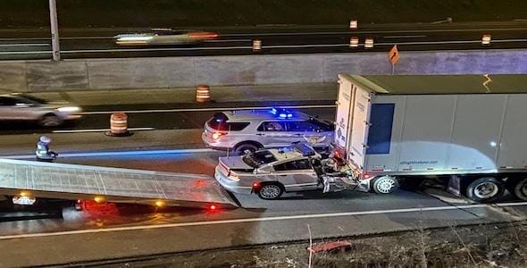 A car is wedged under the rear of a semi-truck on the highway, illustrating one of the common types of truck accidents. Emergency vehicles with flashing lights surround the scene, while a tow truck is poised in front. In the background, traffic snakes past cautiously.