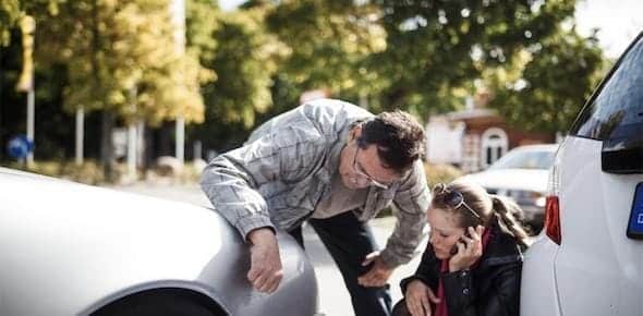 A man inspects a car's fender while a woman kneels nearby, discussing vehicle damage on her phone. Two cars are parked close together on a tree-lined street, possibly considering Michigan Mini Tort claims.