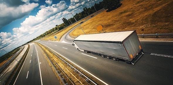 A truck, backed by Michigan Truck Cargo Accident Lawyers, cruises along a curved highway surrounded by fields and forests under a partly cloudy sky.