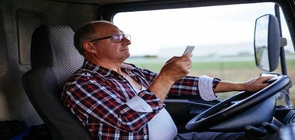 A man in a plaid shirt and glasses sits in a vehicle, holding a mobile phone in one hand and grasping the steering wheel with the other, perhaps contemplating contacting his truck accident lawyer for advice.