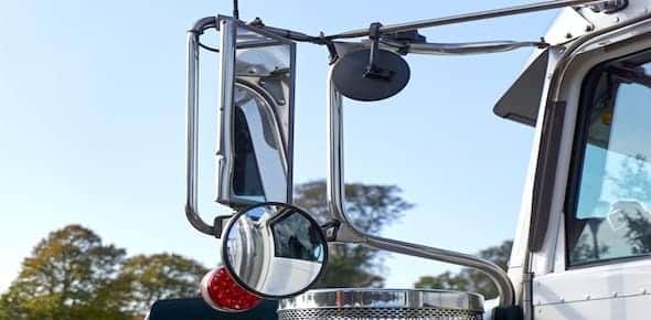 Close-up of a truck's side mirrors and a red signal light, highlighting potential blind spots, with trees and a clear blue sky in the background.
