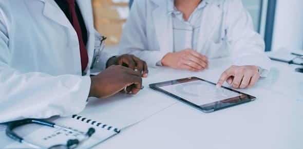 Two doctors in white coats discuss the importance of medical care information on a tablet at a table with a notepad and stethoscope nearby.
