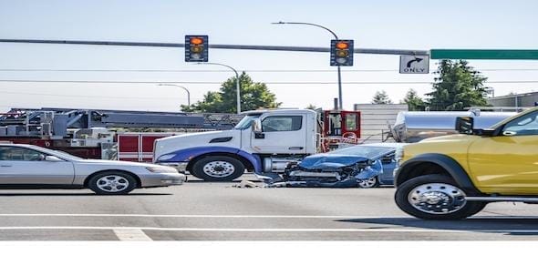 A truck crash occurred at an intersection with traffic lights, involving a blue car and a white truck. Amidst the chaos, other vehicles navigated around, their drivers possibly sipping coffee and rubbernecking at the scene.