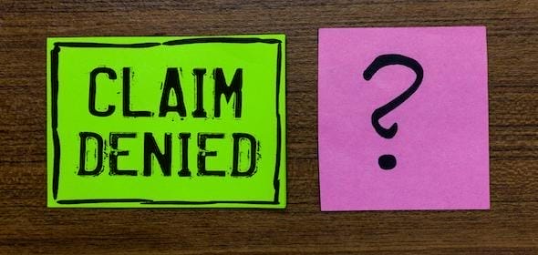 A green note reads "CLAIM DENIED" next to a pink note with a question mark on a wooden surface, echoing the frustration of an auto accident claim in Michigan involving an aggravated pre-existing medical condition.