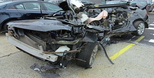 Severely damaged car with a crumpled front and exposed interior, likely the result of a harsh accident, parked on a street near other vehicles.