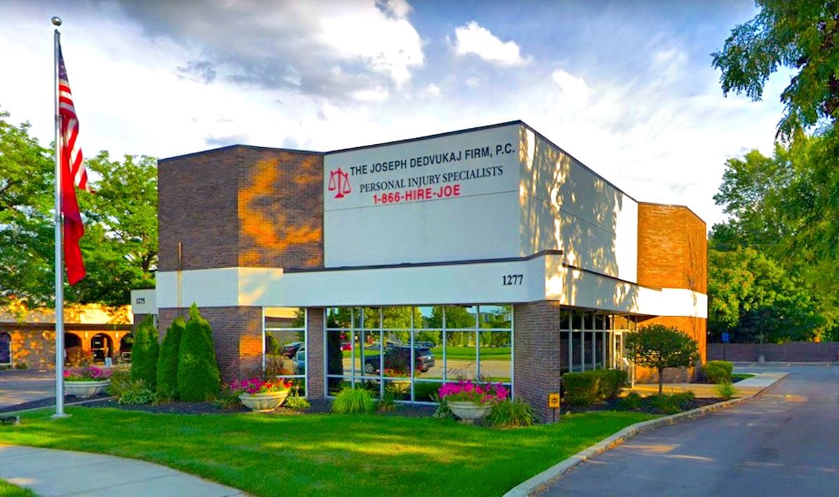 A law firm's building with the name "The Joseph Dedvukaj Firm, P.C." displayed. An American flag is on the left, and green landscaping is in front.