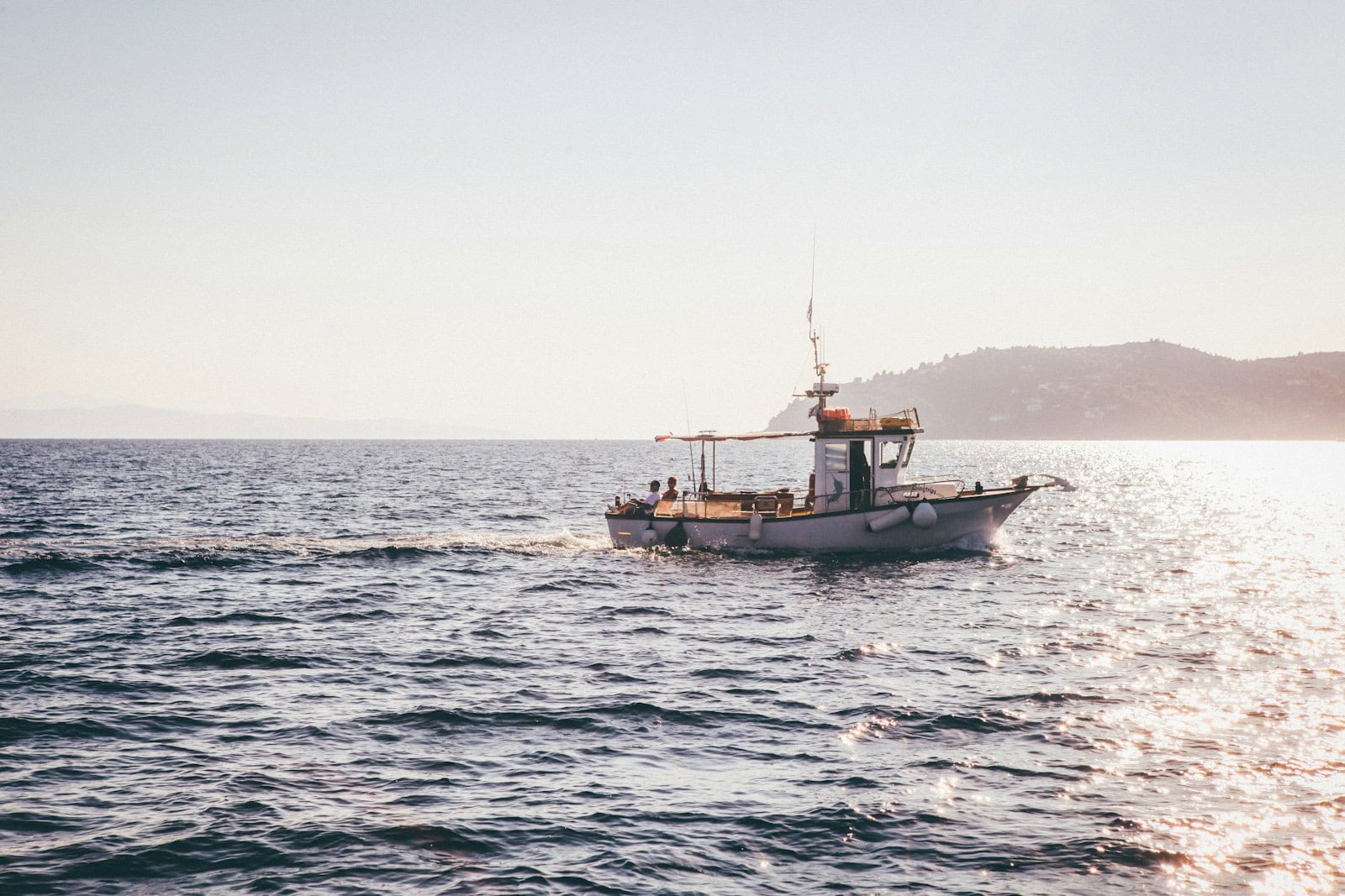 A small fishing boat glides peacefully across a calm sea under a clear sky, with a distant shoreline and rolling hills framing the picturesque scene.