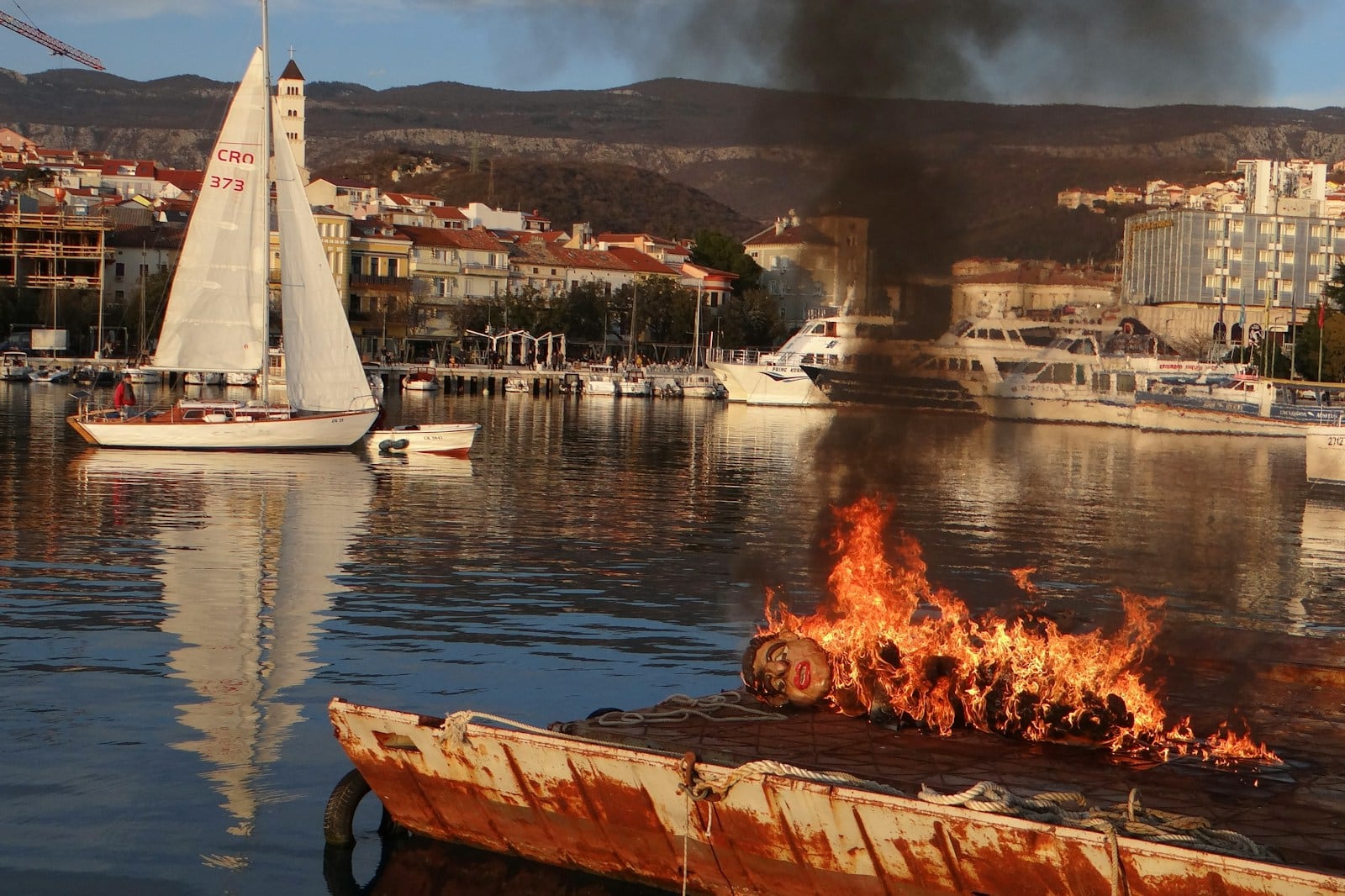 In the harbor, a burning raft with objects ablaze casts flickering light near a sailboat. Smoke billows upward, painting a dramatic scene against the backdrop of buildings and mountains—a stark reminder of how swiftly an accident can unfold amidst the idyllic setting graced by a distant yacht.